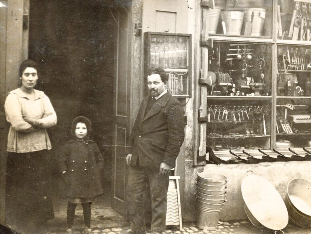 A black-and-white photo of a woman, a young girl, and a man standing outside a hardware store. The shop displays metal buckets, tools, and household items in the window and on the ground.