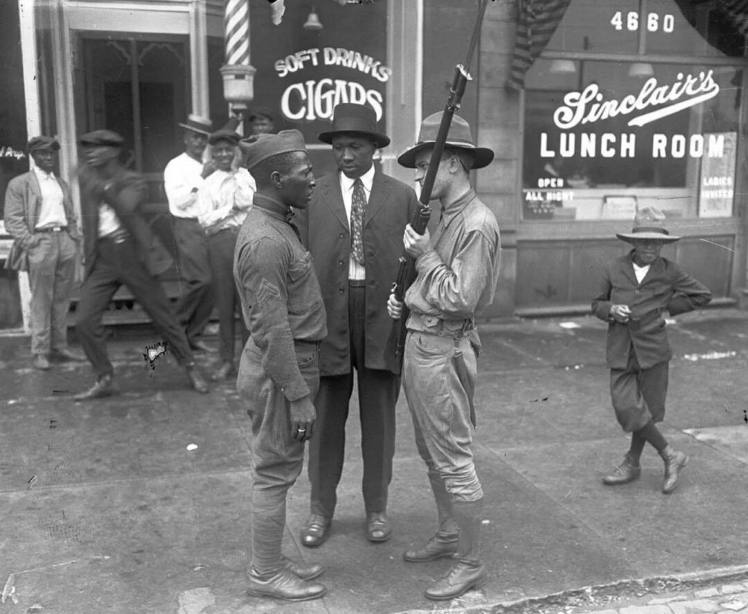 A Black soldier stands face-to-face with a white soldier holding a rifle, outside Sinclair’s Lunch Room, while a group of men and a boy look on from the sidewalk in an early 20th-century urban setting.