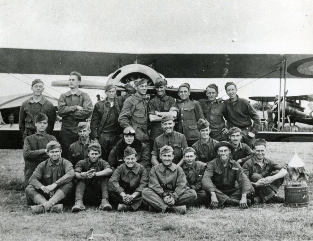 A group of soldiers in uniform pose for a photo in front of a vintage military airplane on a grassy field. The men are smiling, some standing and some sitting cross-legged, with the aircraft's propeller visible behind them.