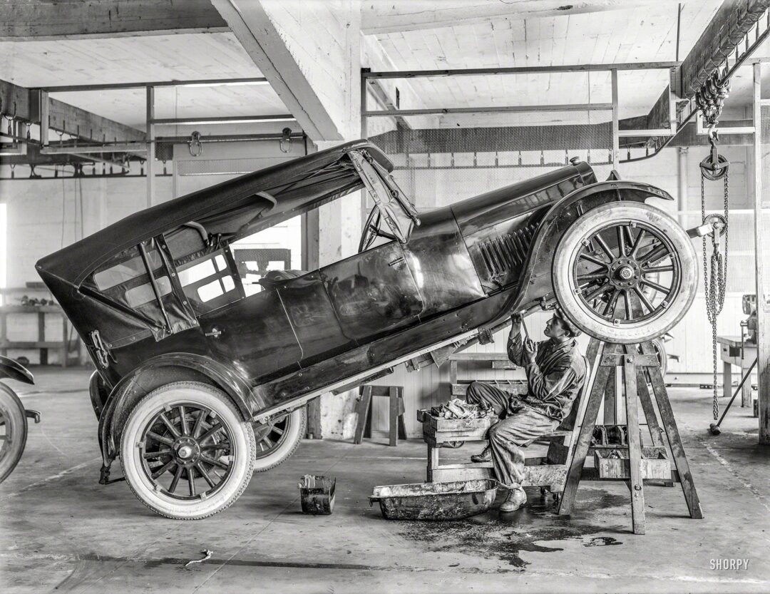 A mechanic in overalls works underneath a vintage car elevated at an angle on supports in a spacious, industrial garage. The car’s front wheels are off the ground, and tools are scattered nearby.