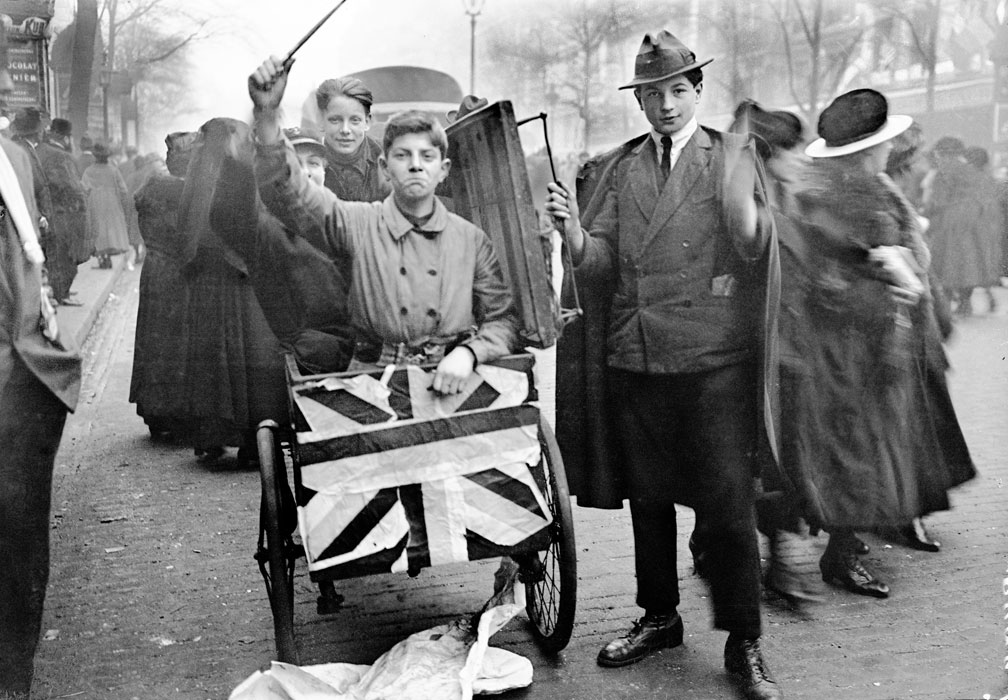 A young person sits in a cart draped with a British flag, raising a stick, while another stands beside them on a busy street with blurred pedestrians, suggesting a parade or public celebration.