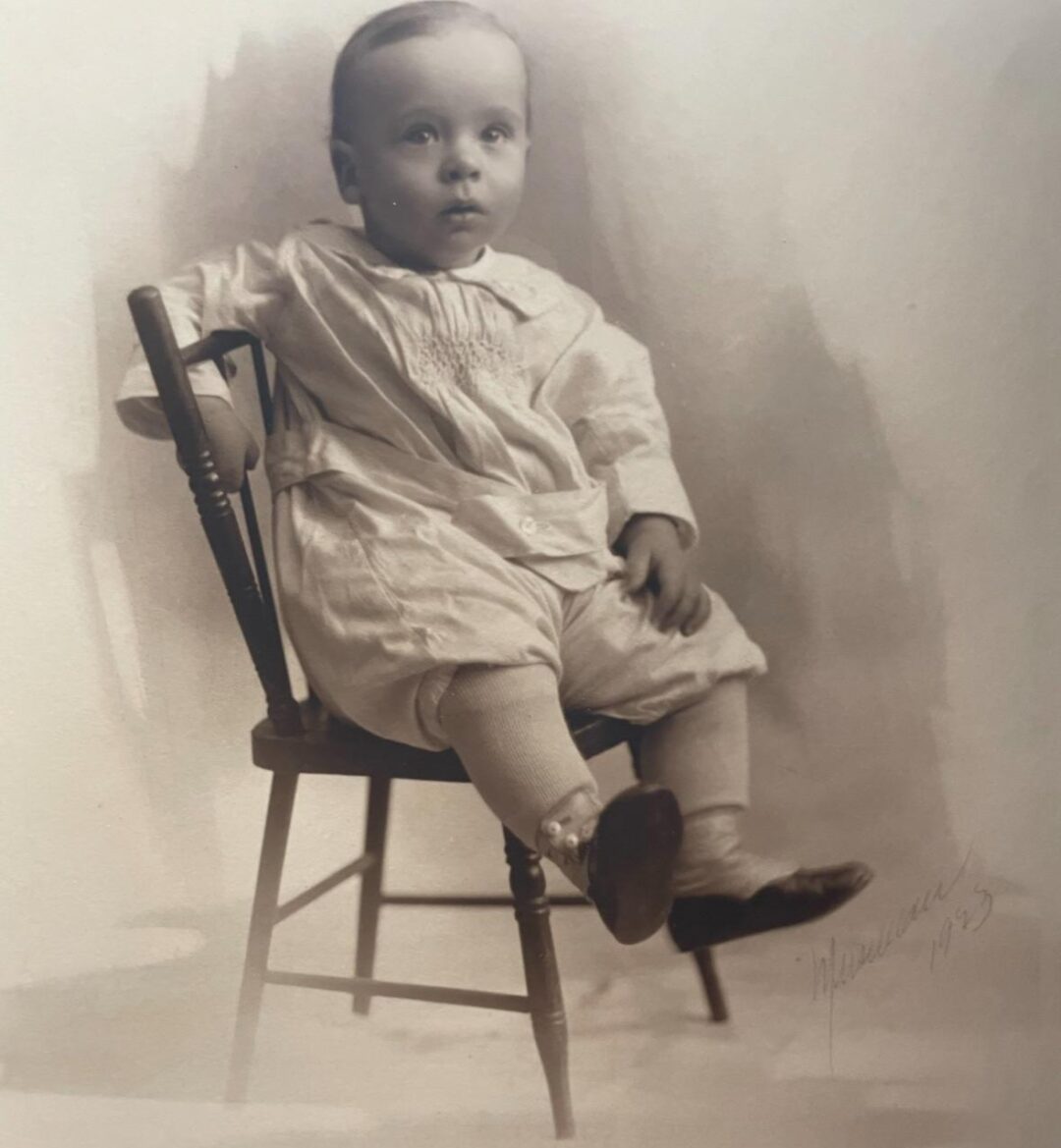 A sepia-toned photograph of a young child sitting sideways on a wooden chair, dressed in a light, old-fashioned outfit with leggings, looking slightly upward. The image is signed "Marmuseel 1923" in the bottom right corner.