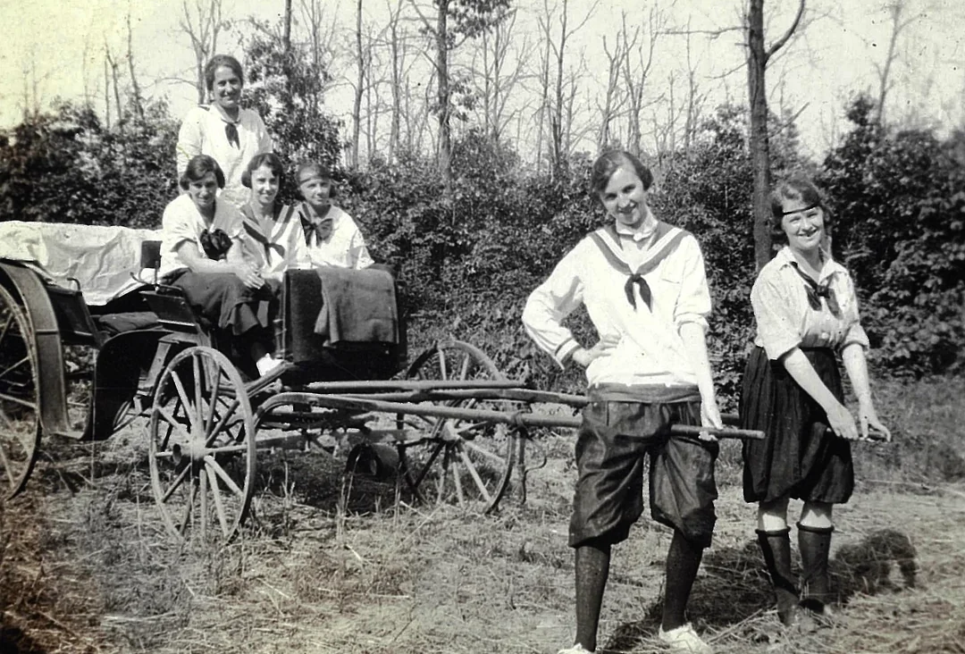 Six young women in vintage clothing pose outdoors; four sit on a horse-drawn wagon, while two stand in front holding the wagon shafts. Trees and bushes fill the background, and the scene has an old-fashioned, nostalgic feel.