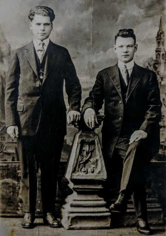 Two young men in early 20th-century suits pose for a formal studio portrait. One stands with a hand on a decorative pedestal, while the other sits with one leg raised on the pedestal. A painted backdrop is behind them.