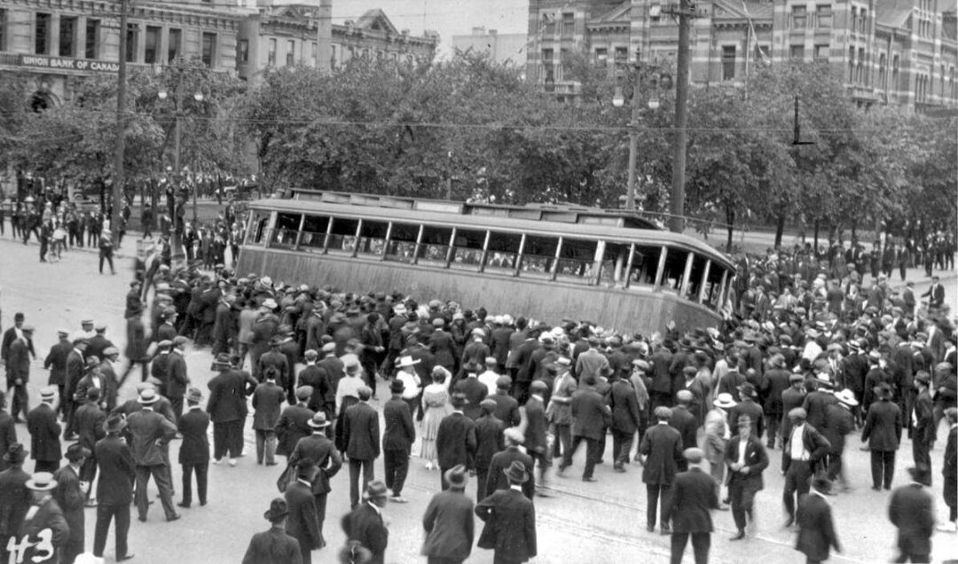 A large crowd of people in early 20th-century attire gathers around a tilted streetcar in a city square, with trees and historic buildings in the background.