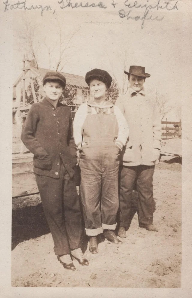 Three people stand outdoors in vintage clothing. Two wear hats and jackets, while the person in the center wears a cap and overalls. Bare trees and a wooden structure are visible in the background. Handwritten names appear at the top.