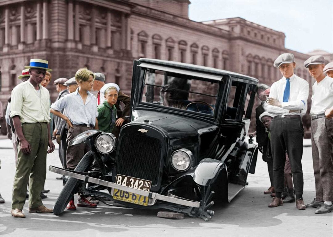 A group of boys and young men gather around a vintage car with a broken front wheel on a city street in front of a large building. The scene appears to be from the early 20th century.