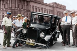 A group of boys and young men gather around a vintage car with a broken front wheel on a city street in front of a large building. The scene appears to be from the early 20th century.