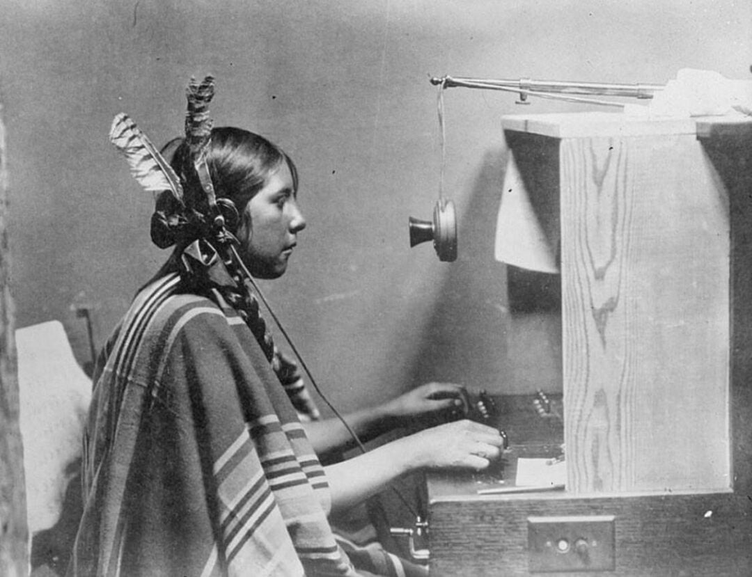 A young Indigenous woman wearing a feathered headdress and a striped blanket uses an old-fashioned telephone switchboard, sitting in profile at a wooden desk.
