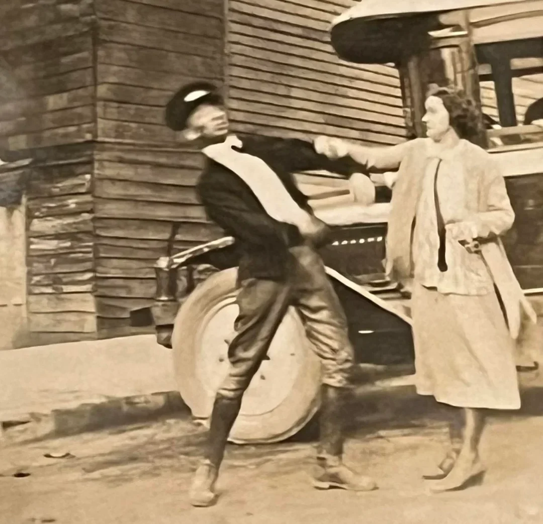 A vintage photo shows a man playfully leaning back as if hit, while a smiling woman stands beside him. They are in front of an old wooden building and an early automobile. Both wear 1920s-style clothing.