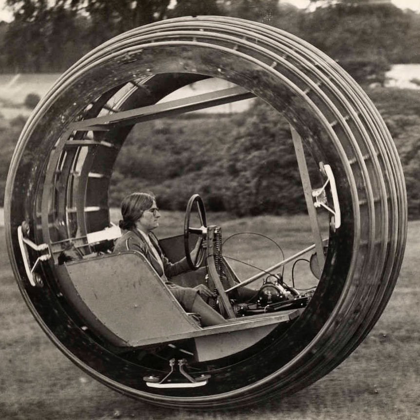 A person sits inside a large, single-wheel vehicle, operating the controls. The unusual design resembles a giant ring with the driver and machinery positioned in its center, outdoors with trees and grass in the background.