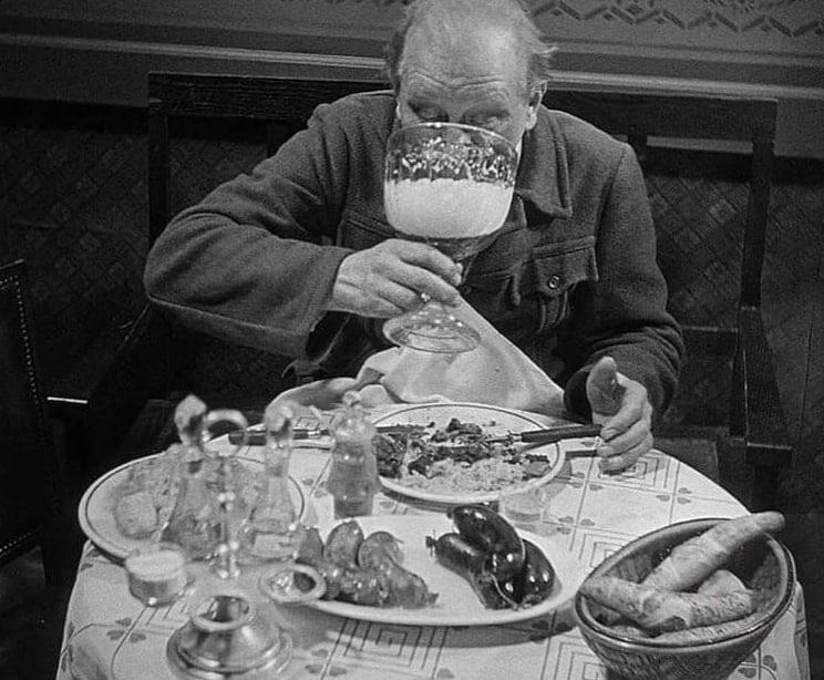 A man sits at a table covered with plates of food, holding and drinking from an oversized goblet filled with a foamy beverage, partially obscuring his face. The table has bread, utensils, and various dishes.
