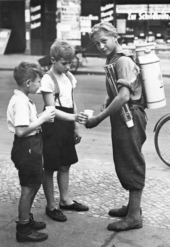 Three boys stand on a city sidewalk. One boy, wearing a container on his back and carrying cups, pours a drink for another boy, while a third boy waits. Buildings and a bicycle are visible in the background.