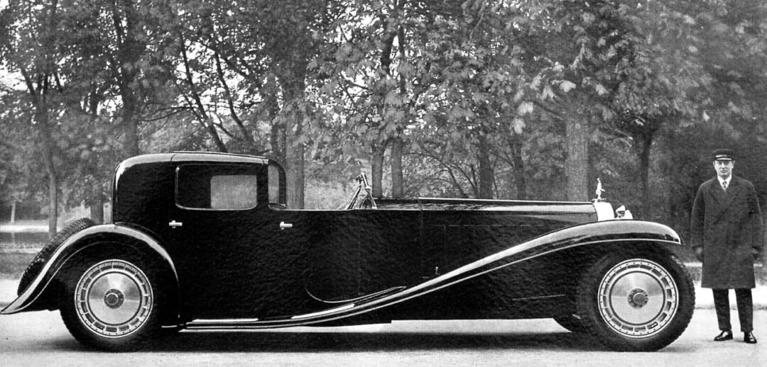 A vintage black luxury car with sweeping fenders is parked outdoors near leafy trees. A man in a suit and hat stands beside the car, highlighting its impressive length and classic design.
