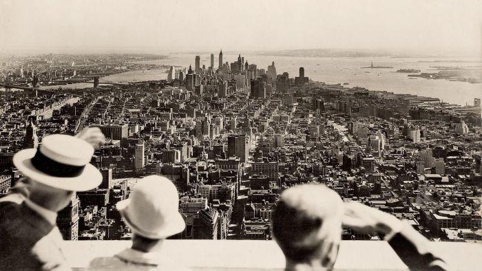 Three people in hats look out over the New York City skyline from a high vantage point in the 1930s, with skyscrapers, rivers, and the Statue of Liberty visible in the distance.