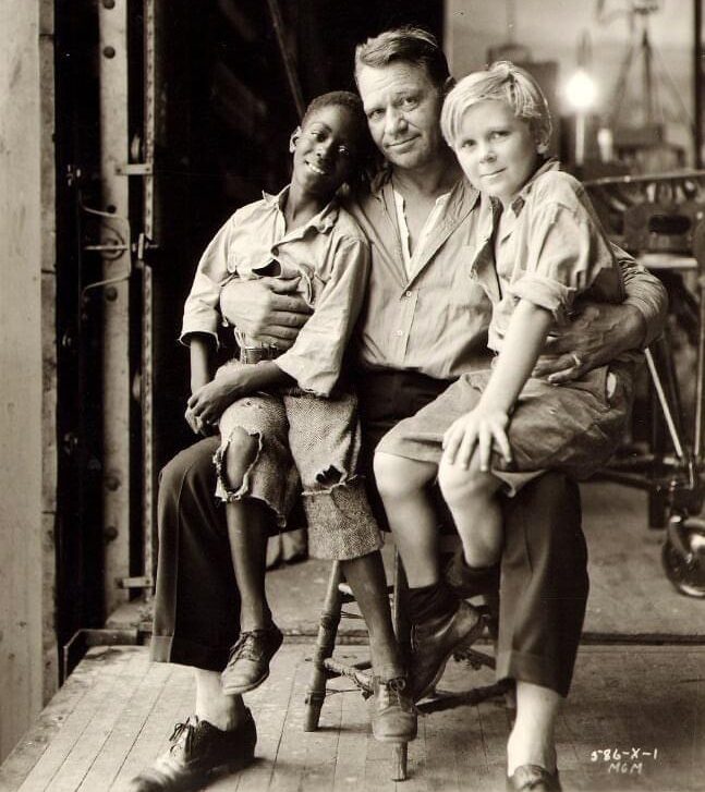 A man sits on a chair with two boys on his lap—one Black, wearing a ripped shirt and shorts, and one white, dressed in light-colored clothes. They all look toward the camera in an industrial-looking setting.