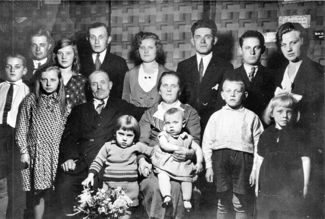 A black and white vintage photo of a large family, with men, women, and children posing indoors in formal and casual attire. Some adults are seated in front, with children and other family members standing behind them.