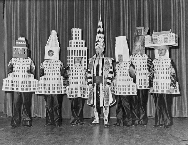 A group of people dressed in costumes resembling famous New York City buildings stand on stage with a central figure in a decorative outfit; all are in front of a curtain. The image is in black and white.