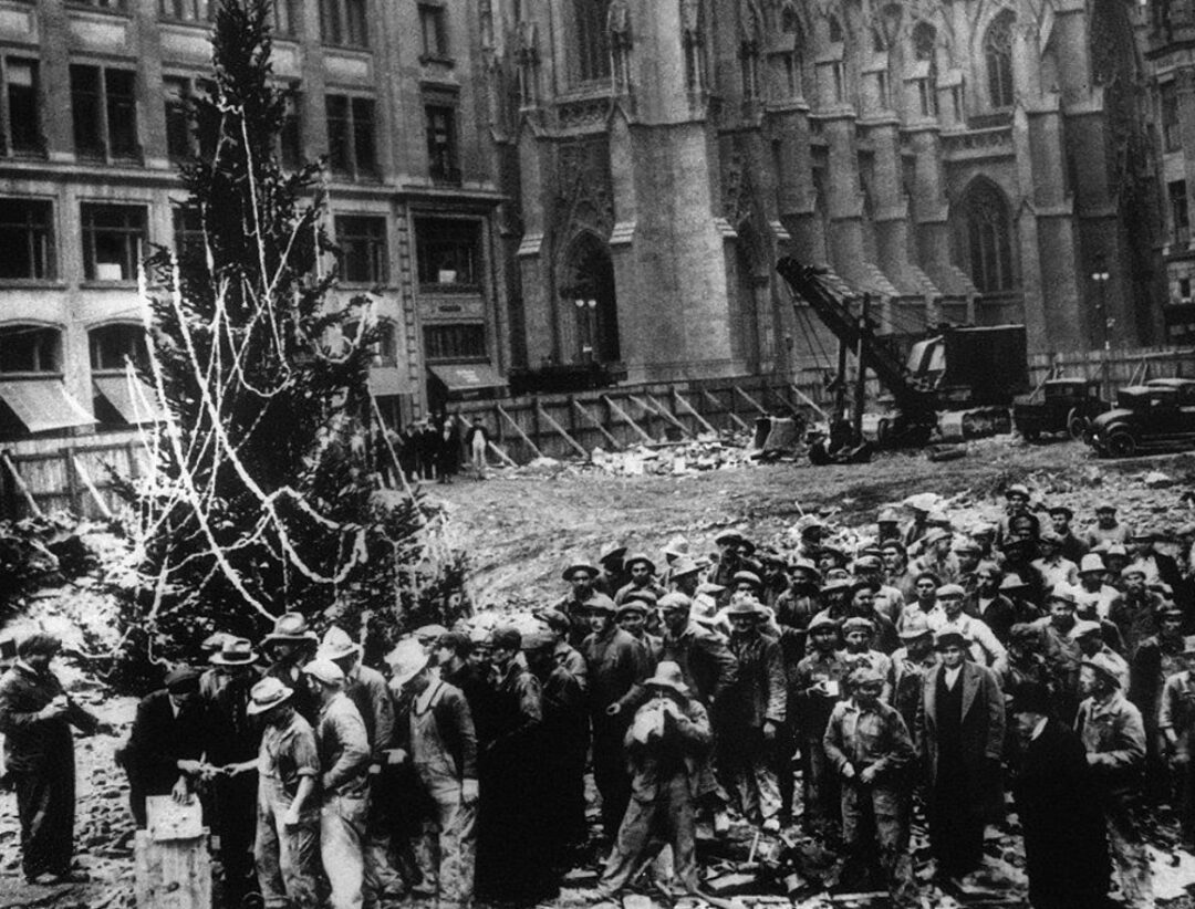 A large group of construction workers gathers around a decorated Christmas tree at a building site in a city, with tall buildings and construction equipment in the background. The scene appears to be from the early 20th century.
