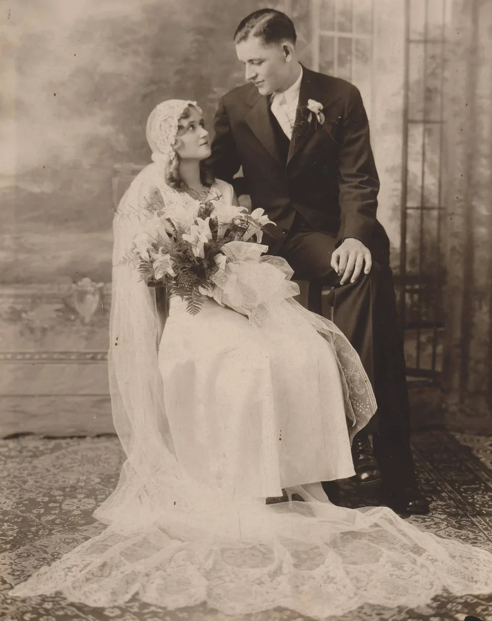 A vintage black and white photo shows a bride in a lace dress and veil holding a bouquet, seated beside a groom in a dark suit. They look at each other lovingly, posing against a painted studio backdrop.