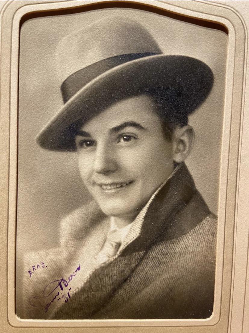 A vintage sepia photo of a young man smiling, wearing a wide-brimmed hat, a suit, and a thick collared coat. The portrait is framed, and there is handwriting and a signature visible in the bottom corner.