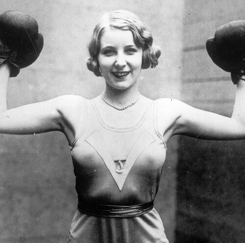 A smiling woman in vintage athletic attire poses with her arms raised, wearing boxing gloves. The black-and-white photo appears to be from the early 20th century.