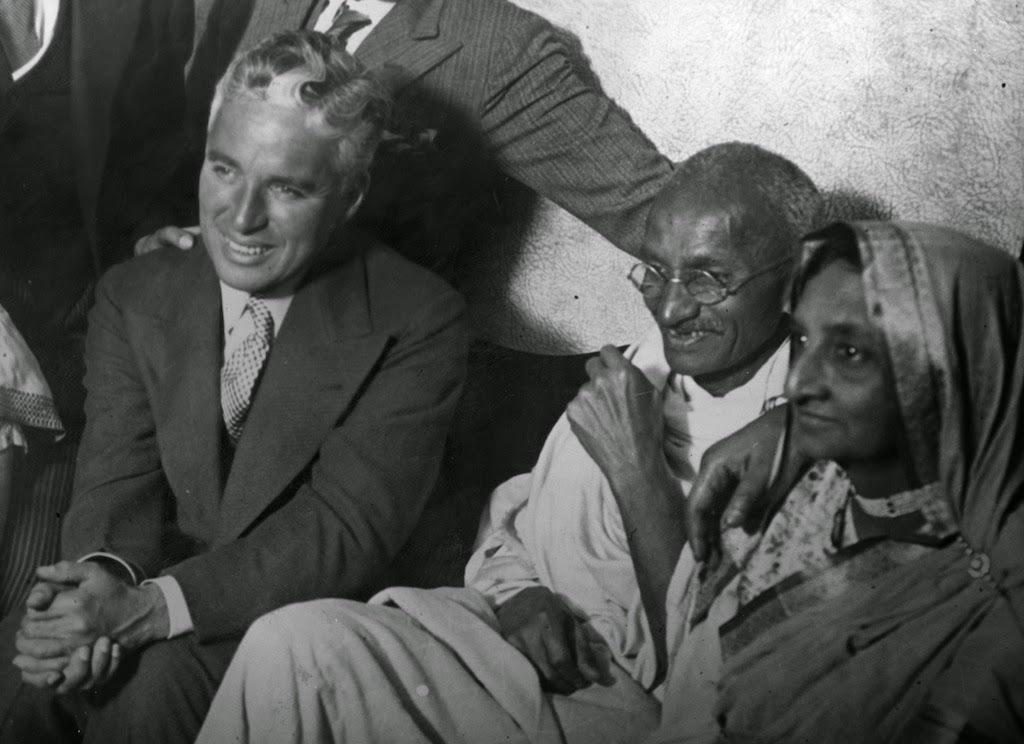 Three people sit closely together; a man in a suit smiles on the left, while a man in glasses and simple white clothing and a woman in a sari sit beside him, both looking ahead.