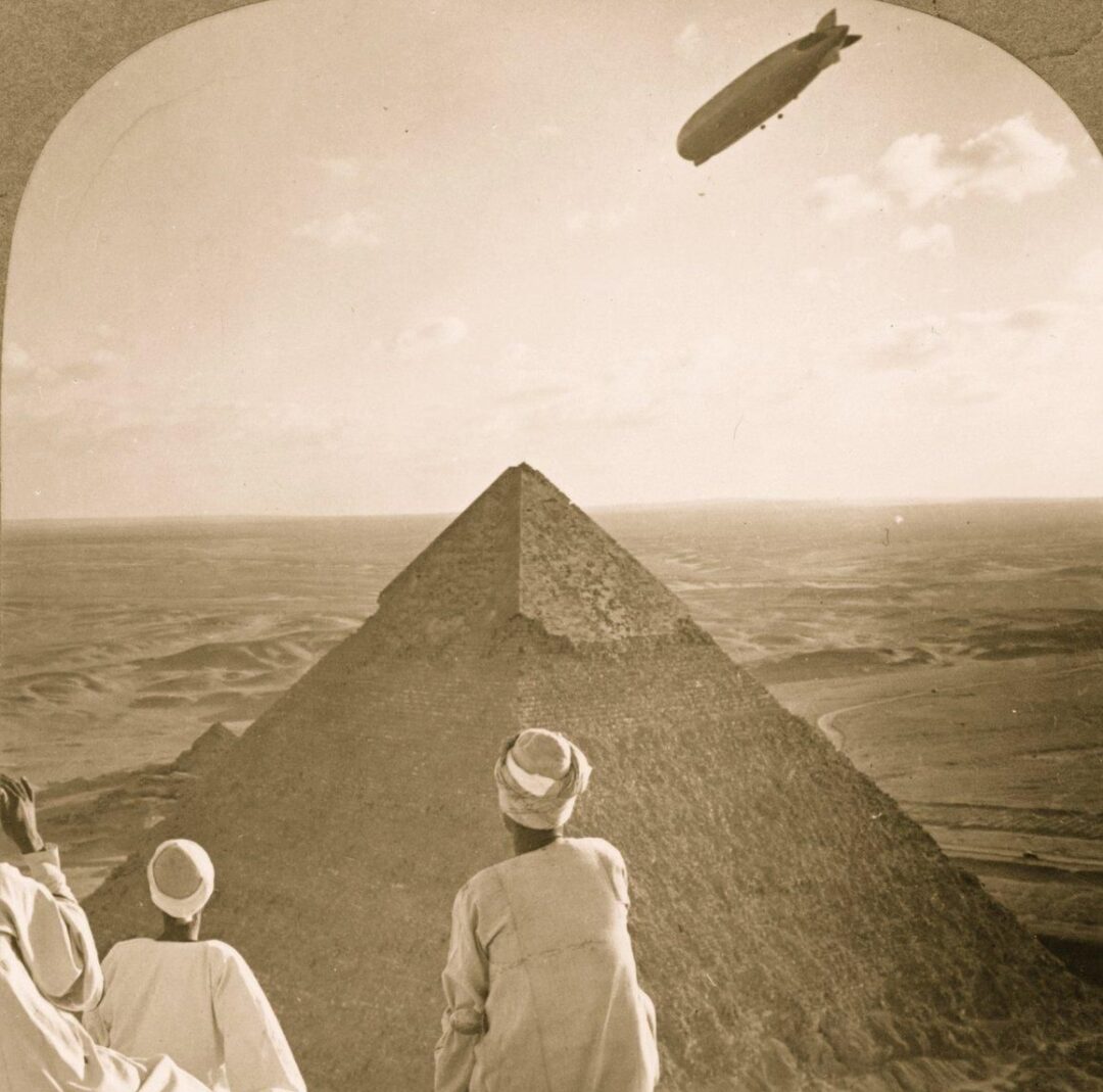 Three people in traditional clothing sit atop the Great Pyramid of Giza, looking out over the desert as a large zeppelin floats in the sky above the horizon.