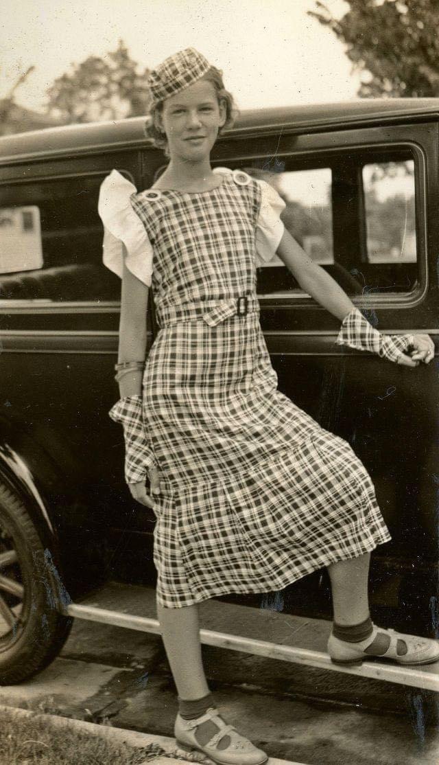 A young girl poses by a vintage car, wearing a plaid dress with puffed sleeves, matching hat and gloves, and strap shoes. She smiles at the camera with one foot on the car’s running board. The photo appears old and sepia-toned.