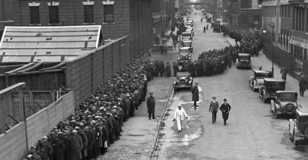 A long line of people, mostly men in coats and hats, waits along a city street beside a brick wall. Several old-fashioned cars are parked, and a few people, including uniformed officers, walk on the street. The scene is from the early 20th century.