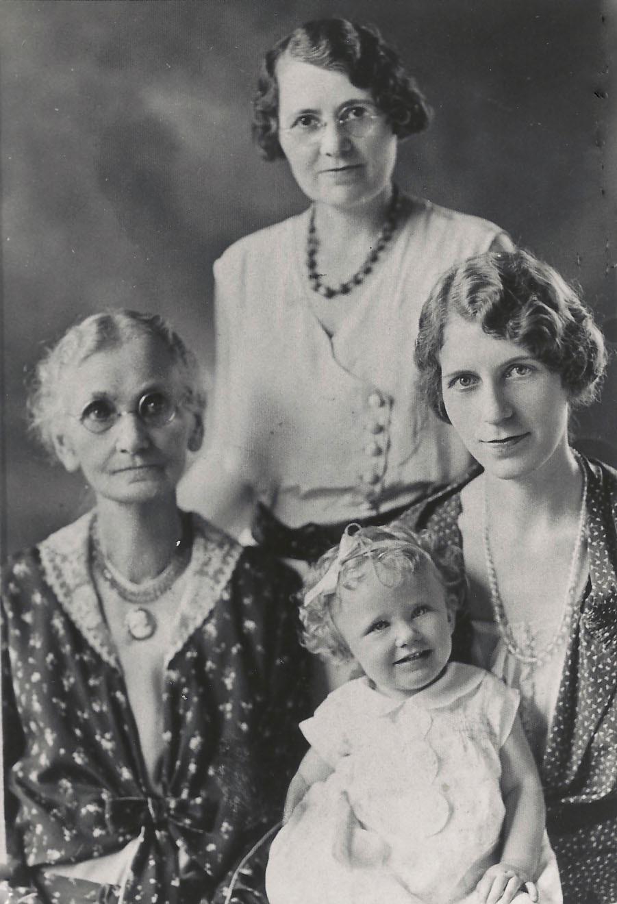 Four generations of women pose together for a black-and-white portrait: an elderly woman with glasses, a middle-aged woman standing behind, a younger woman, and a smiling toddler in front.