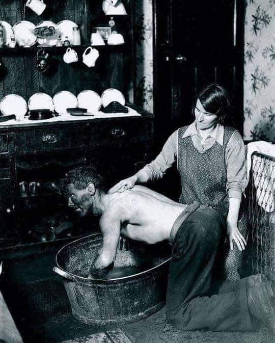 A woman kneels beside a shirtless man who is bathing in a metal tub; she is washing his back in a modest, old-fashioned kitchen with dishes and cups on shelves in the background.