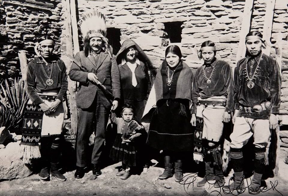 A black-and-white photo of six adults and one child standing in front of a stone building. Some adults wear traditional Native American attire and jewelry; one man wears a suit and feathered headdress.