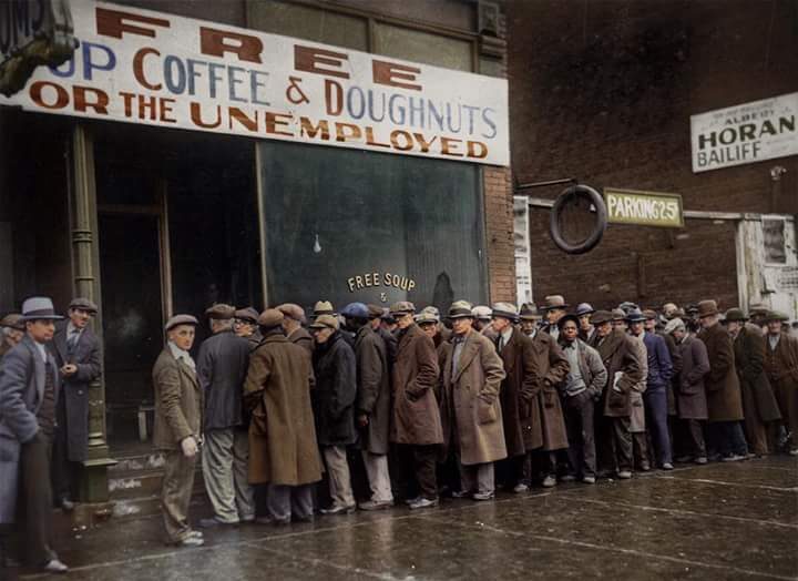 A long line of men in coats and hats wait outside a storefront offering free soup, coffee, and doughnuts for the unemployed. The sign above the door advertises the free food, reflecting the hardship of the Great Depression era.