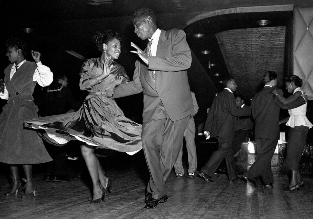 Black and white photo of elegantly dressed couples swing dancing energetically on a wooden dance floor in a lively, mid-20th century club setting. The lead couple smiles joyfully while caught mid-move.
