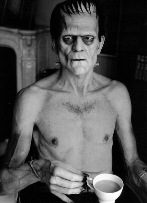 A person in Frankenstein’s monster makeup and costume sits shirtless at a table, holding a white teacup. The scene is in black and white, and the background shows part of a fireplace and shelves.
