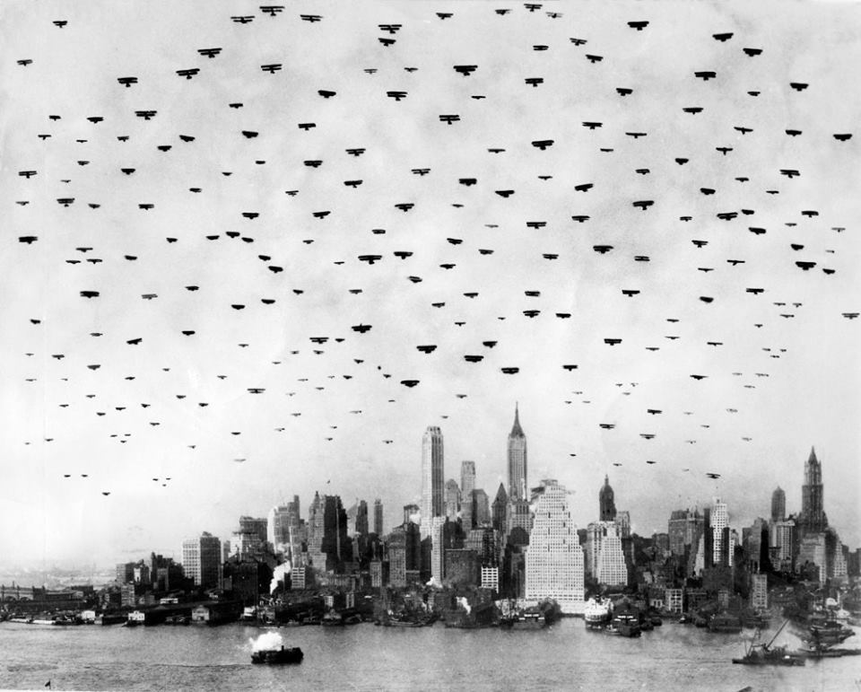 Dozens of vintage airplanes fly in formation over the New York City skyline, with iconic skyscrapers visible below and a boat on the river in the foreground. The image is in black and white.