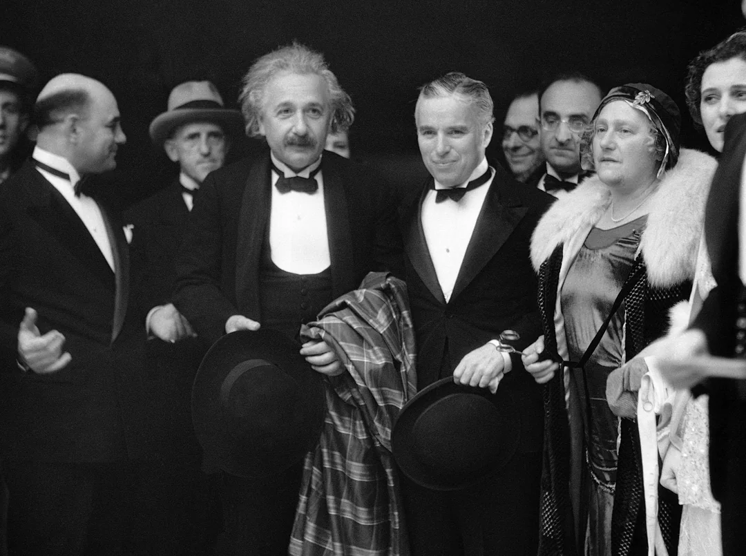 A black-and-white photo of Albert Einstein and Charlie Chaplin standing together in formal attire, surrounded by other people in evening wear, smiling and posing for the camera.