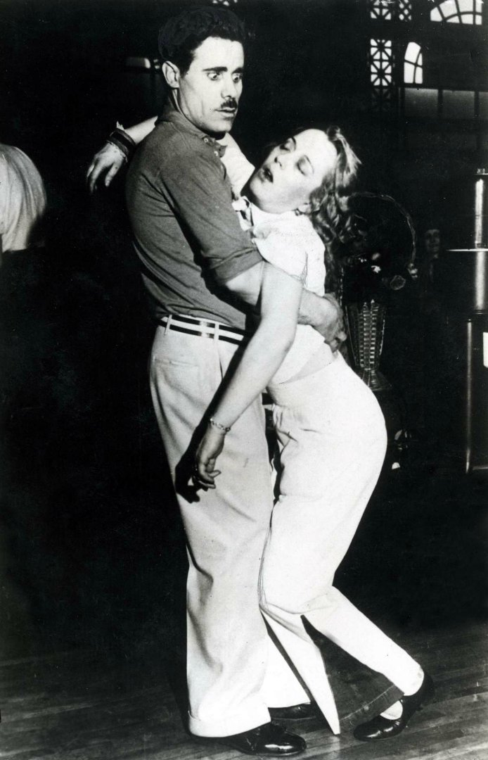 A man and woman dance closely; the man holds the woman firmly as she leans back with eyes closed, appearing dramatic or faint. Both wear vintage-style clothing in a dimly lit room.
