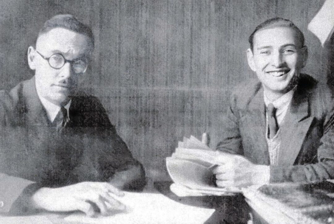 Two men in suits sit at a desk with papers. The man on the left wears round glasses and looks serious, while the man on the right smiles, holding a stack of documents. Both are in a black-and-white photo.