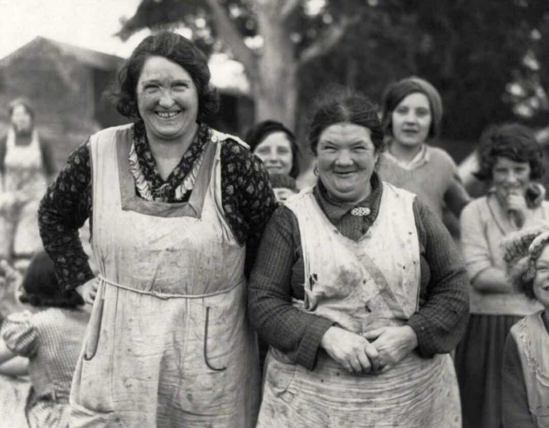 Two women wearing aprons stand smiling in the foreground, their clothes and aprons appear dirty. Several women and children stand behind them outdoors, all looking toward the camera. The photo is black and white.