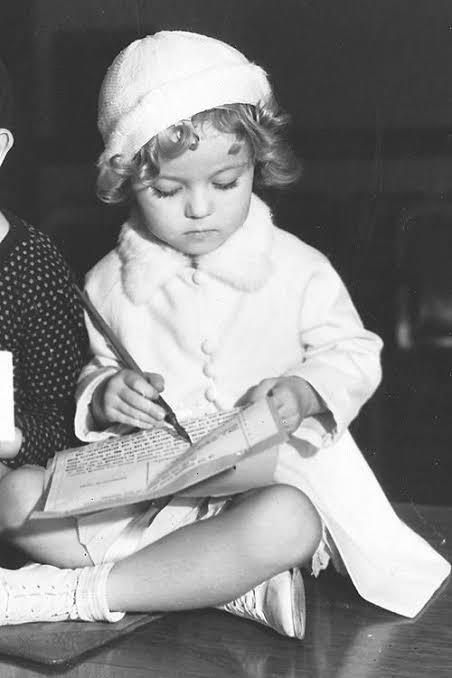 A young girl with curly hair, wearing a white coat and hat, sits on the floor and writes in a notebook with a pencil, looking down with a focused expression.