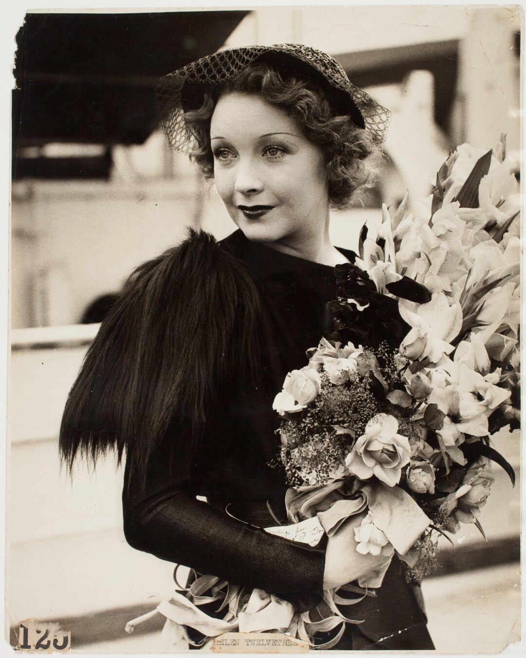 A woman wearing a dark dress with fur accents and a netted hat holds a large bouquet of flowers. She has short, wavy hair and is looking to the side with a slight smile. The photo is black and white.