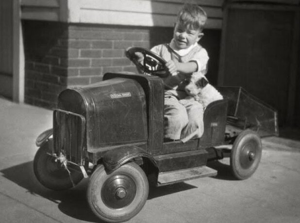 A young boy sits in a vintage pedal car with a small dog beside him, outdoors near a brick building. The image is black and white, giving it a nostalgic, old-fashioned feel.