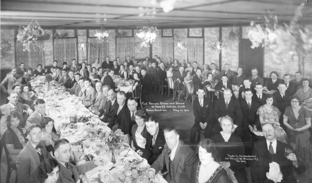 A large group of men and women in formal attire sit at long banquet tables set with food and flowers, attending a social club dinner in an ornately decorated room, circa 1920s.