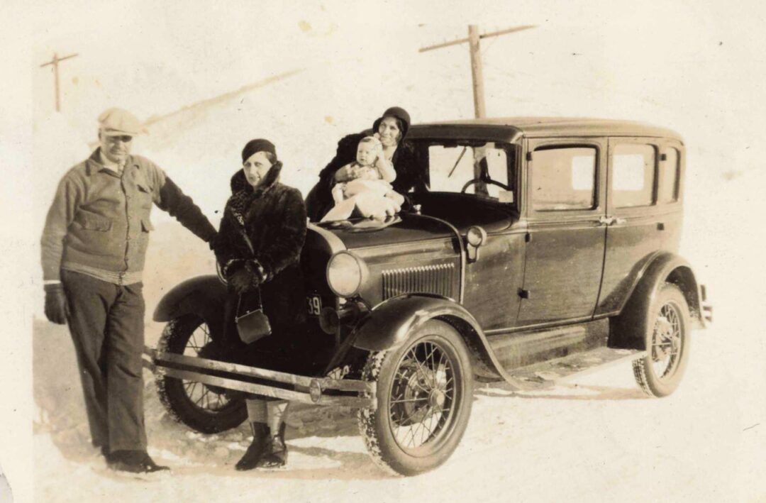 A sepia-toned photo of three adults and a child posing with a vintage car on a snowy road. Two women and the child sit on the car, while a man stands beside it. Wooden utility poles are visible in the background.