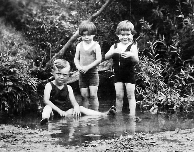 Three young children in old-fashioned swimsuits stand and sit at the edge of a shallow, muddy pond surrounded by lush plants. Two children smile, while one sits in the water with a serious expression.
