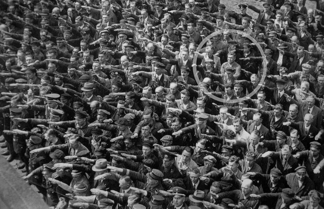 A large crowd of men stands closely packed, many giving the Nazi salute. In the center, one man stands with his arms crossed, not participating. A white circle highlights him, emphasizing his refusal among the group.