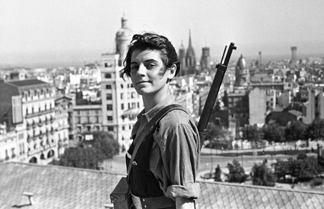 A young woman with short dark hair stands on a rooftop, wearing overalls and carrying a rifle on her back. Behind her is a cityscape with historic buildings and a cathedral, under a clear sky.
