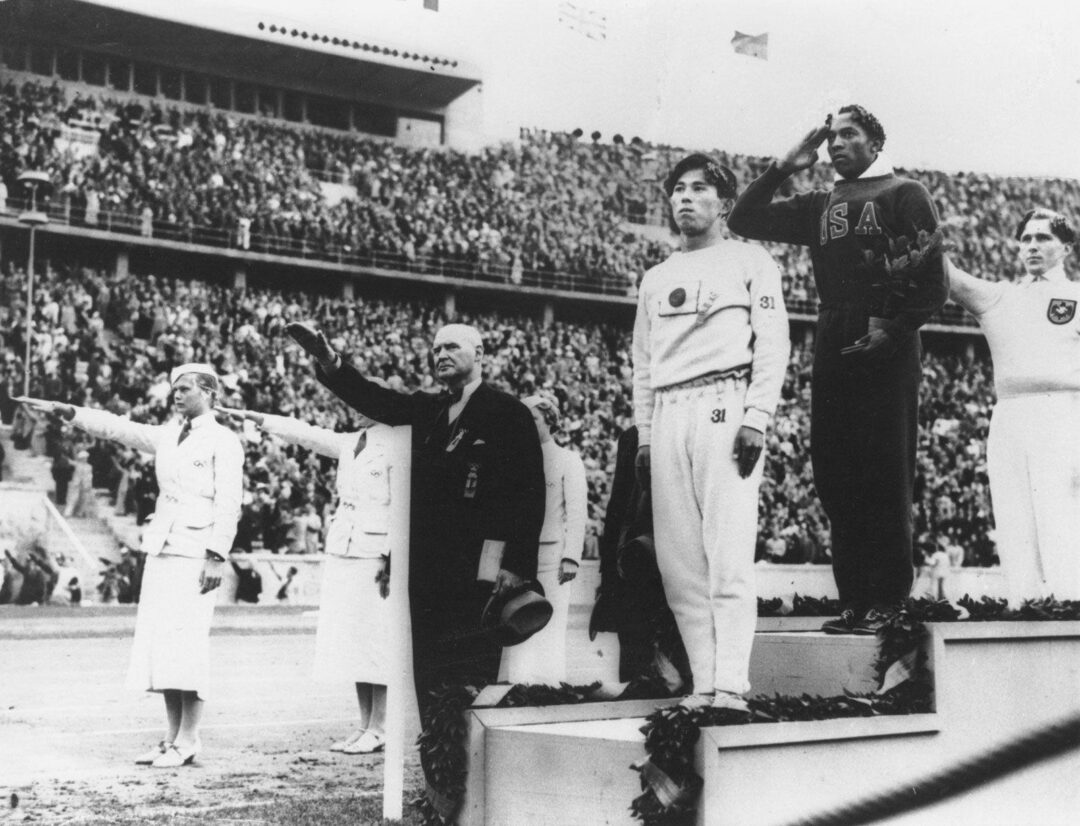 Three athletes stand on a podium at a stadium during a medal ceremony. The gold medalist, in a dark tracksuit, raises his right hand in a salute, while others nearby, including officials, perform a different salute.